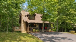 A log cabin with a stone chimney, a porch with red chairs, and a well-maintained lawn.