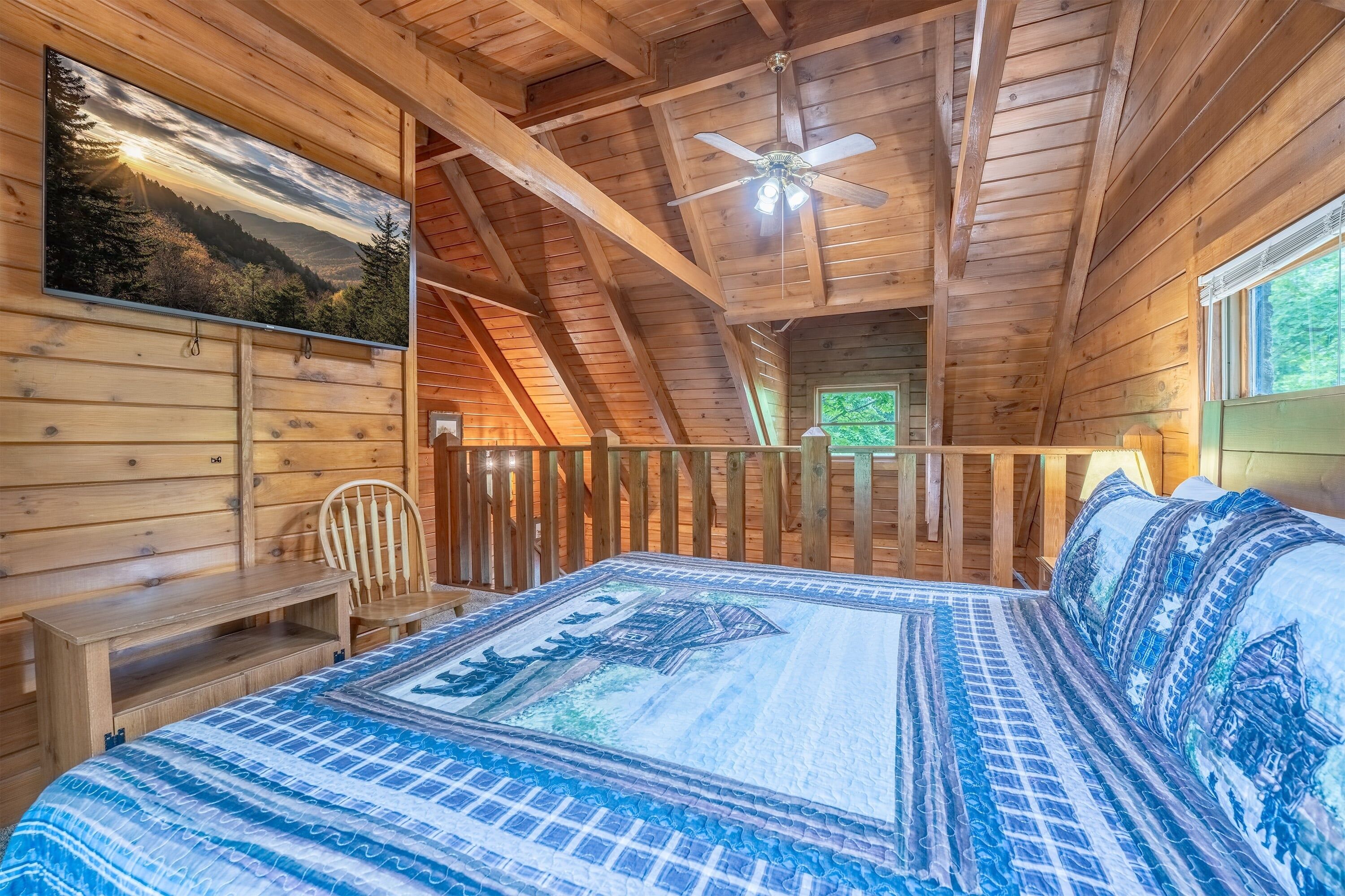 A wooden cabin room with a bed, a chair, a desk, a ceiling fan, and a window.