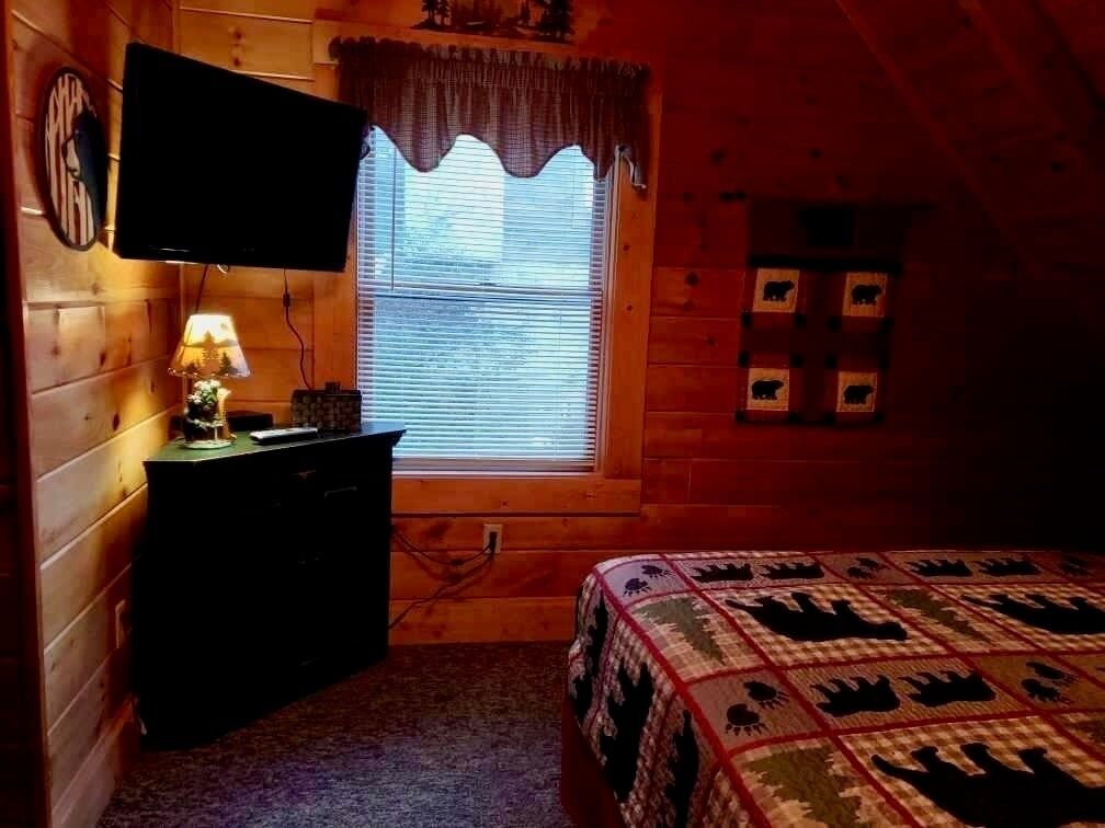 Una habitación cálida tipo cabaña con una cama, un tocador, un televisor y una ventana con persianas.
