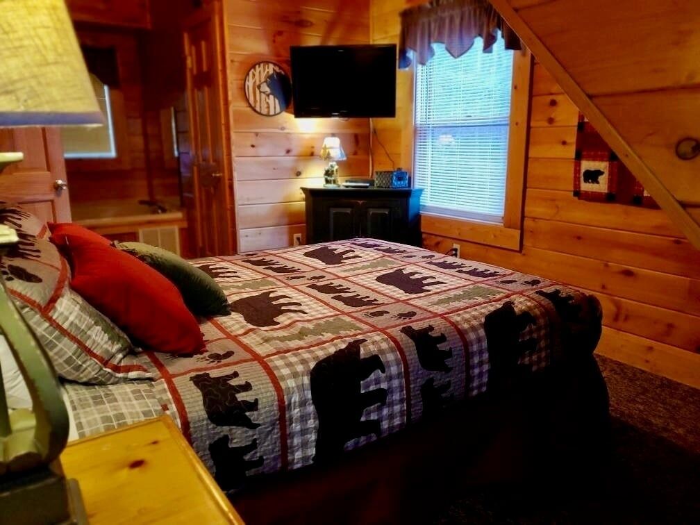 A cozy cabin room with a bed, a television, a window with blinds, and a clock on the wall.