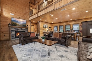 A spacious living room with a fireplace, leather sofas, a dining area, and a high ceiling with wooden beams.