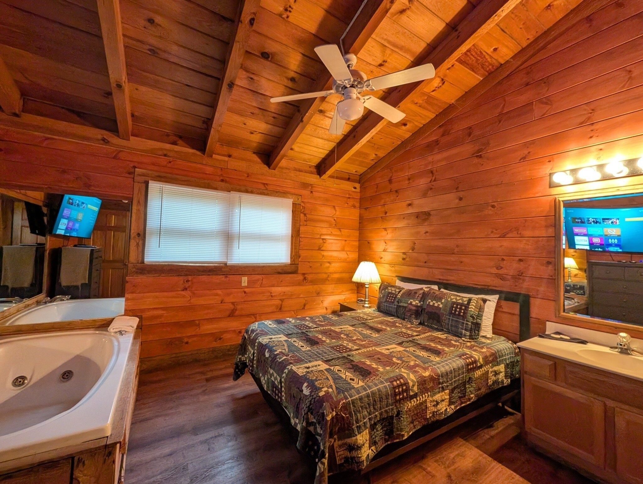 Una habitación de cabaña acogedora con interior de madera, una cama con colcha estampada, un ventilador de techo, un ventanal con persianas, un televisor y una bañera.