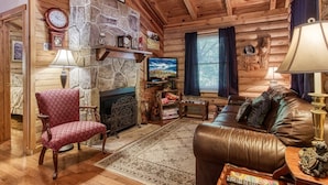 A cozy cabin interior with a stone fireplace, leather sofa, wooden walls, and a television.