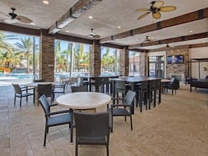 A spacious indoor area with multiple tables and chairs, a pool visible through large windows, and ceiling fans.