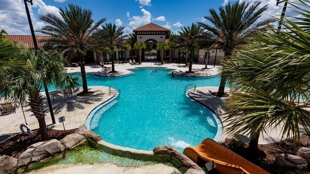 A large outdoor swimming pool with a slide, surrounded by palm trees and a building with a red-tiled roof.
