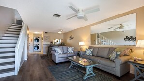A modern living room with a sofa, coffee table, and a dining area. There is a staircase with a handrail and a ceiling fan. - Coastal 2BDRM Townhouse 100 Steps to Beach! (Holmes Beach)
