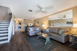 Ein modernes Wohnzimmer mit Sofa, Couchtisch und Essbereich. Eine Treppe mit Geländer und ein Deckenventilator sind ebenfalls vorhanden.