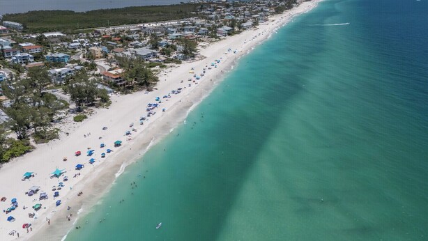 An aerial view of a beach with rows of beach umbrellas and a row of buildings along the shore. - Coastal 2BDRM Townhouse 100 Steps to Beach! (Holmes Beach)