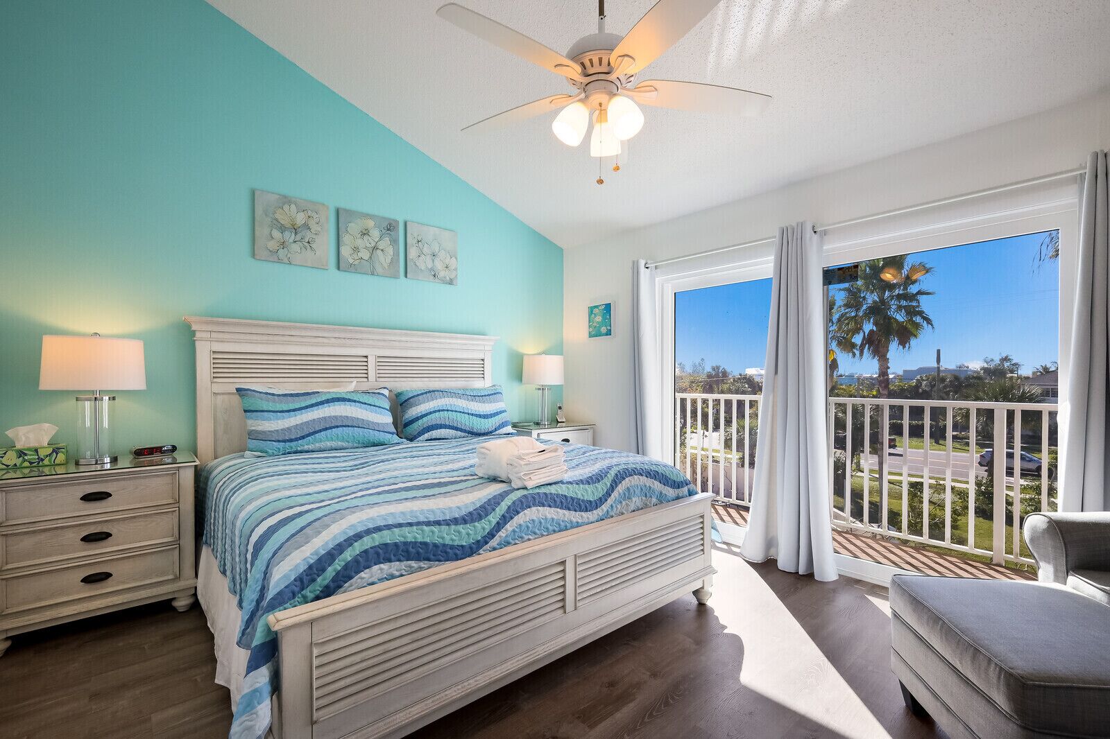 A bedroom with a bed, nightstands, a ceiling fan, and a balcony with a view of palm trees and a road.
