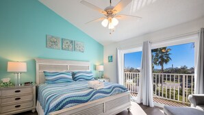 A bedroom with a bed, nightstands, a ceiling fan, and a balcony with a view of palm trees and a road. - Coastal 2BDRM Townhouse 100 Steps to Beach! (Holmes Beach)