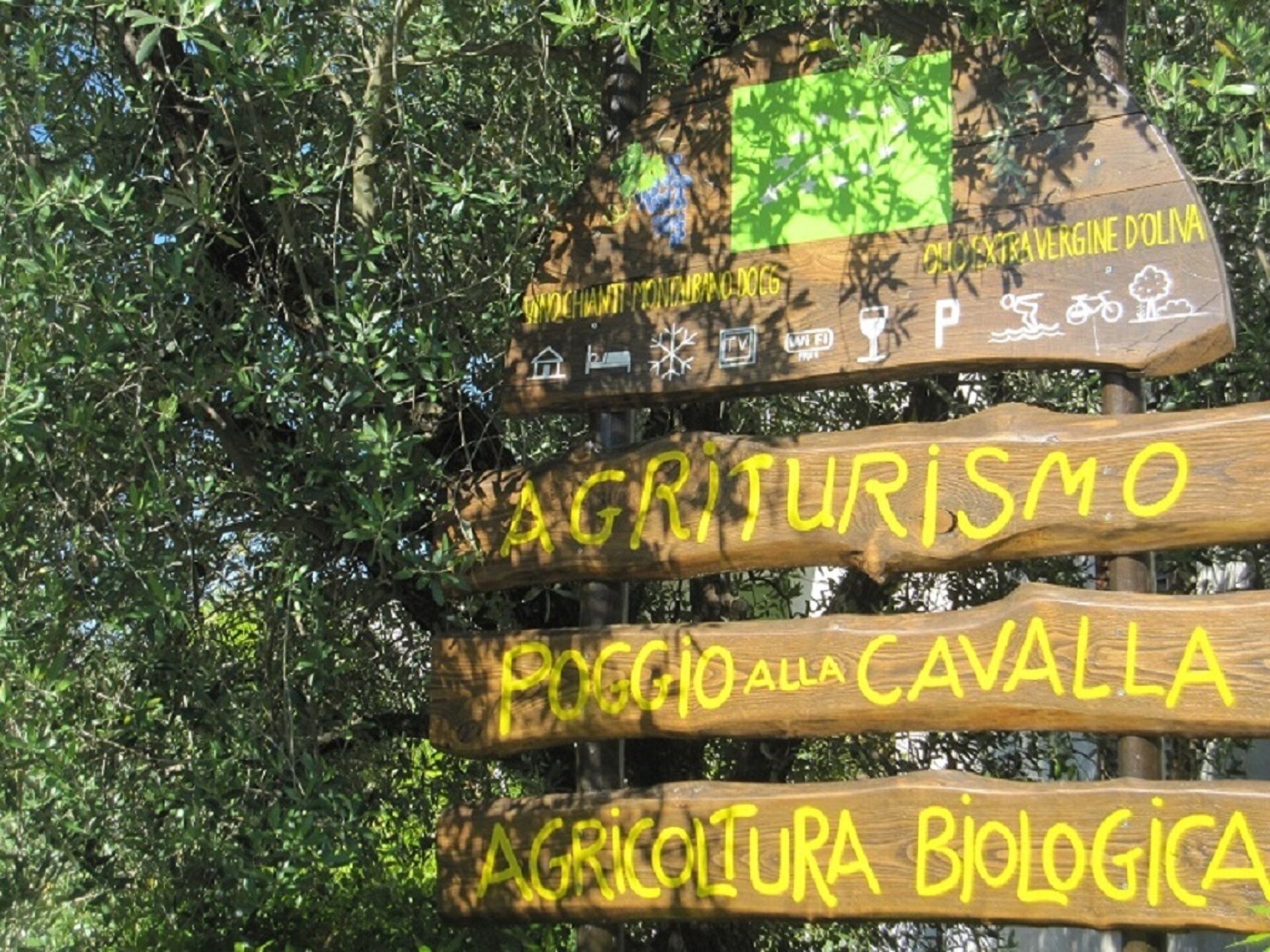 Ein hölzernes Schild mit gelber Schrift, das Wegweiser zu 'Agriturismo', 'Poggio alla Cavalla' und 'Agricoltura Biologica' zeigt.