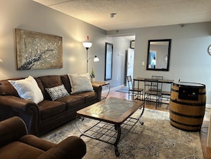 A living room with a brown leather sofa, a coffee table, a dining table with chairs, a wooden barrel, and a painting on the wall. - Gatlinburg Towers Condo Unit 501 - Great Location! Close To Town! 3 Bedroom Condo by RedAwning (Gatlinburg)