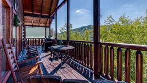A wooden deck with a table and chairs, a grill, and a view of green trees and hills. - Ern850 - Simply The Best - Great Location - Close To All The Action! 4 Bedroom Cabin by RedAwning (Pigeon Forge)
