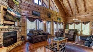 A cozy log cabin interior with a stone fireplace, leather sofas, and a large window overlooking a forest. - Ern850 - Simply The Best - Great Location - Close To All The Action! 4 Bedroom Cabin by RedAwning (Pigeon Forge)