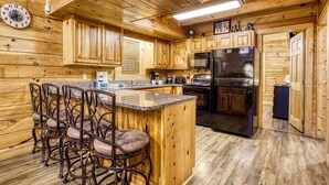 A wooden cabin interior with a kitchen featuring a refrigerator, microwave, and stove. There is a bar area with stools and a clock on the wall.