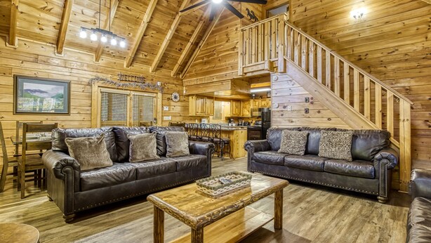 A log cabin interior with two leather sofas, a wooden coffee table, a staircase, and a kitchen area.