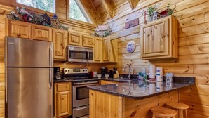 A wooden cabin kitchen with a stainless steel refrigerator, a microwave, an oven, and a sink. There are wooden stools at the island counter. - Er260- Magical Moments Great Location- Close To Town 1 Bedroom Cabin by RedAwning (Pigeon Forge)