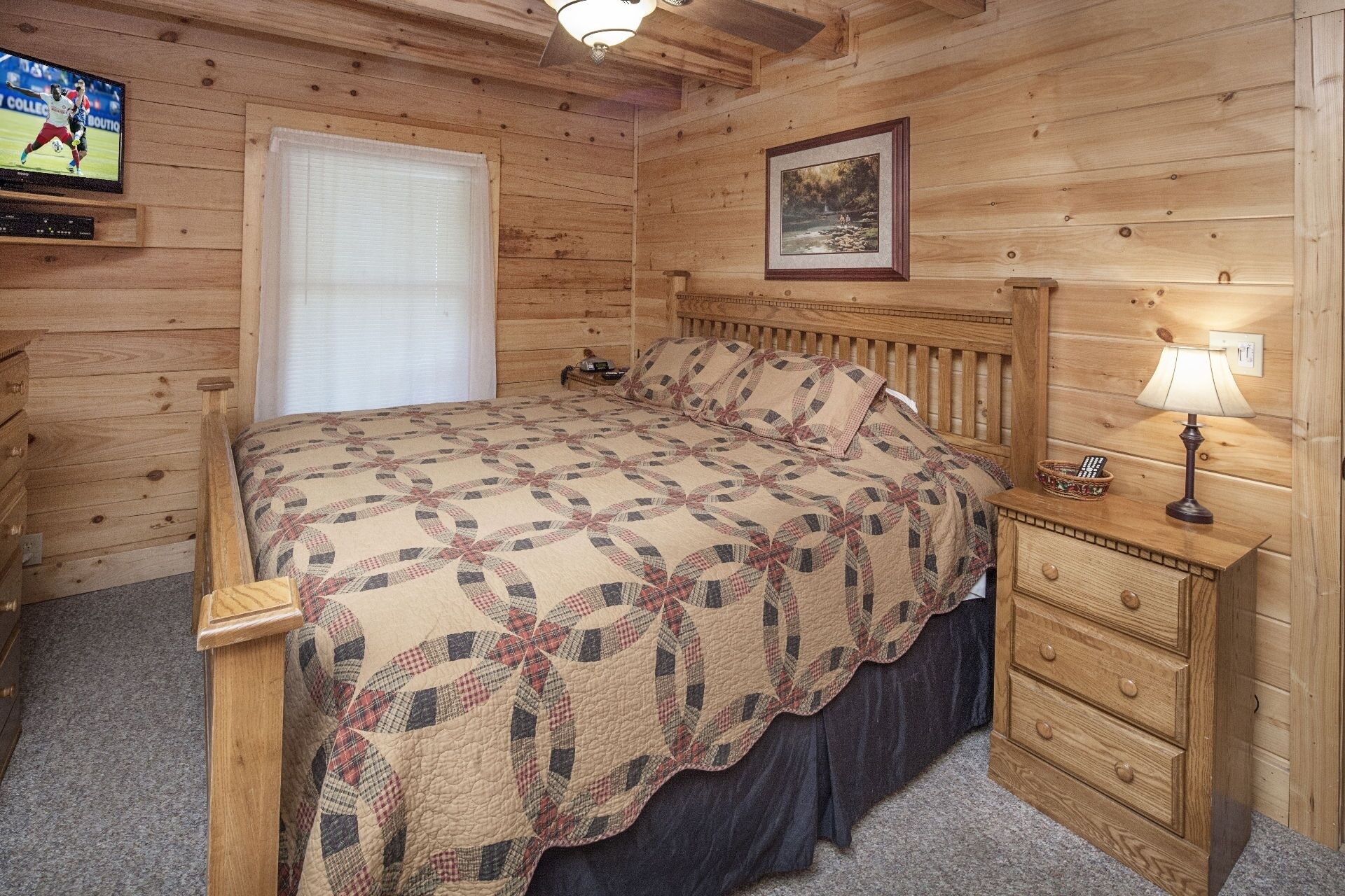 A wooden cabin bedroom with a bed, a nightstand with a lamp, a TV mounted on the wall, and a framed picture above the bed.