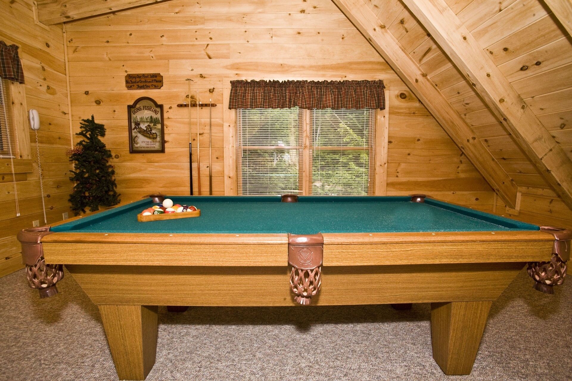A pool table in a wooden room with a sloped ceiling, a Christmas tree, and a window with blinds.