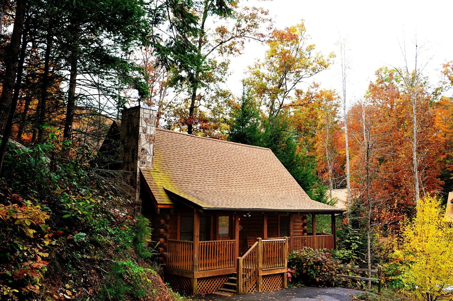 A log cabin with a wooden deck and a chimney, surrounded by autumn foliage.