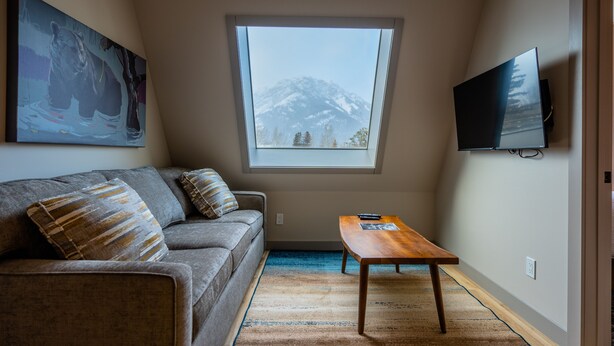 A living room with a sofa, a coffee table, a TV mounted on the wall, and a window with a mountain view. - Superior Two Bedroom Apartment (Banff)