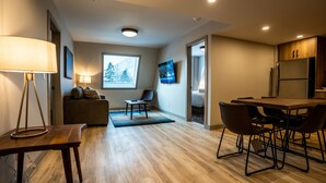 A modern living room with a sofa, coffee table, and dining area with chairs. There is a TV mounted on the wall and a window with a mountain view. - Superior King Two Bedroom Apartment (Banff)