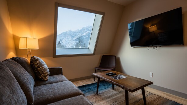 A modern living room with a sofa, a small table, a lamp, a TV, and a window with a mountain view. - Superior King Two Bedroom Apartment (Banff)