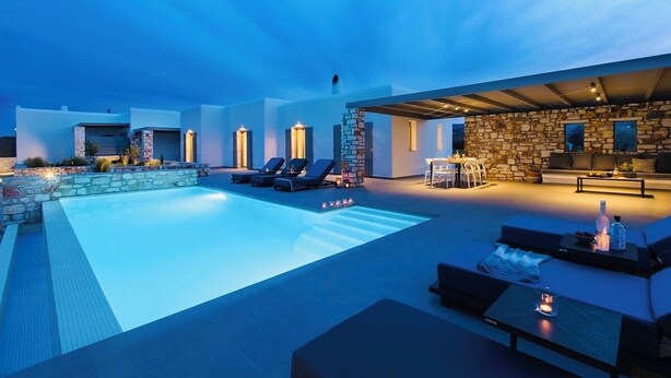 Area piscina esterna moderna con lettini, un muro in pietra e una zona salotto coperta con camino.