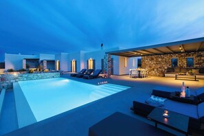 Un área de piscina moderna al aire libre con sillones de descanso, un muro de piedra y una zona de estar cubierta con chimenea.