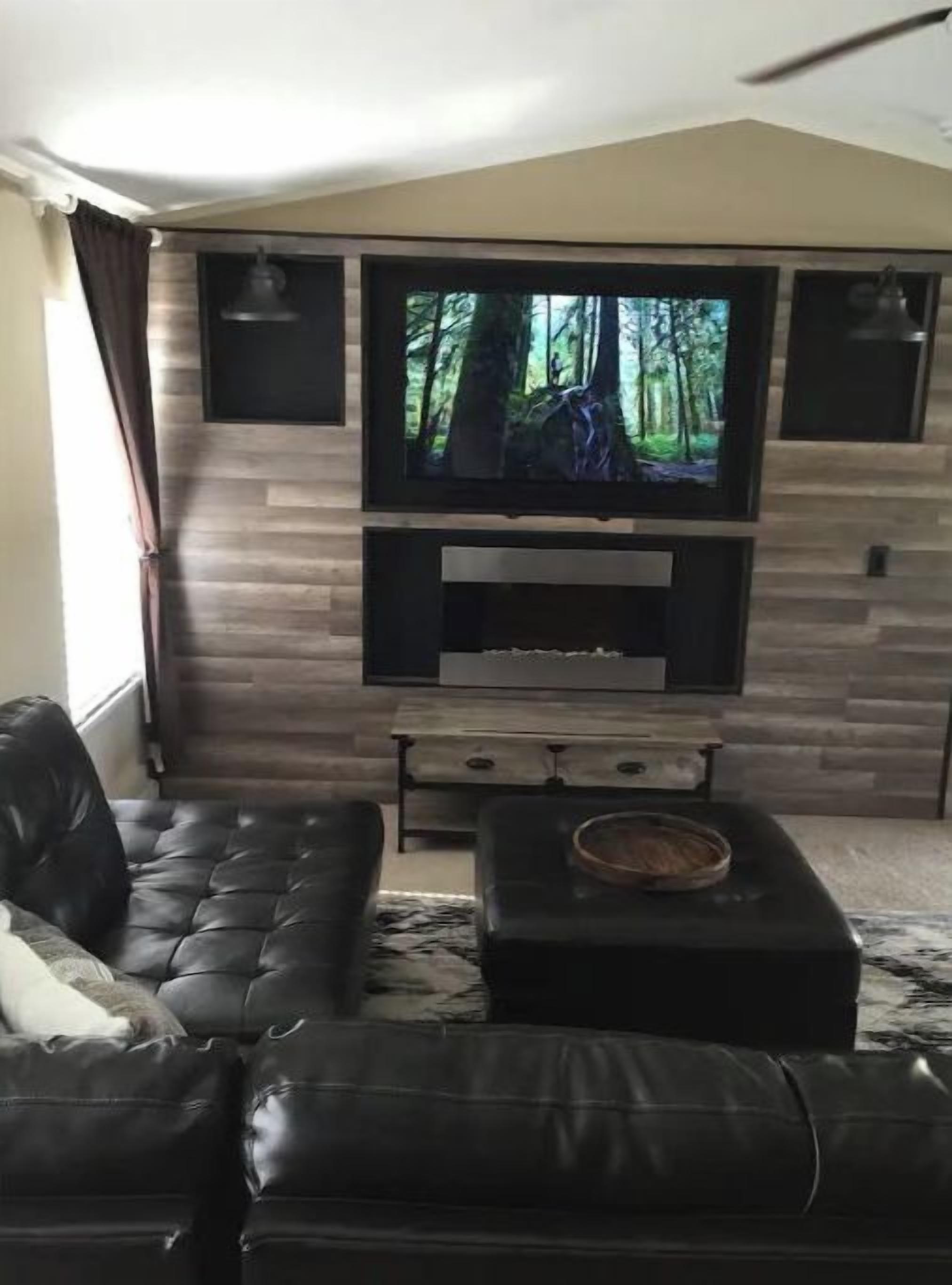 A living room with a black leather sofa, a flat-screen TV mounted on a wooden wall, and a fireplace below.