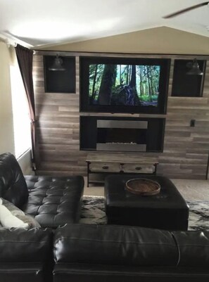 A living room with a black leather sofa, a flat-screen TV mounted on a wooden wall, and a fireplace below.