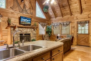 A log cabin interior with a stone fireplace, a stone sink, wooden cabinets, and a television mounted above the fireplace. - Er50 - Creekside - Great Location. Close To All The Action! 2 Bedroom Cabin by RedAwning (Pigeon Forge)