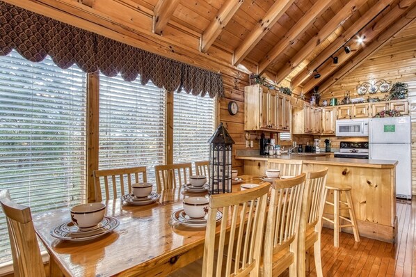 Interior de una cabaña de madera con una mesa de comedor preparada para cuatro personas, sillas de madera, una zona de cocina con armarios de madera, un refrigerador y una ventana con persianas.