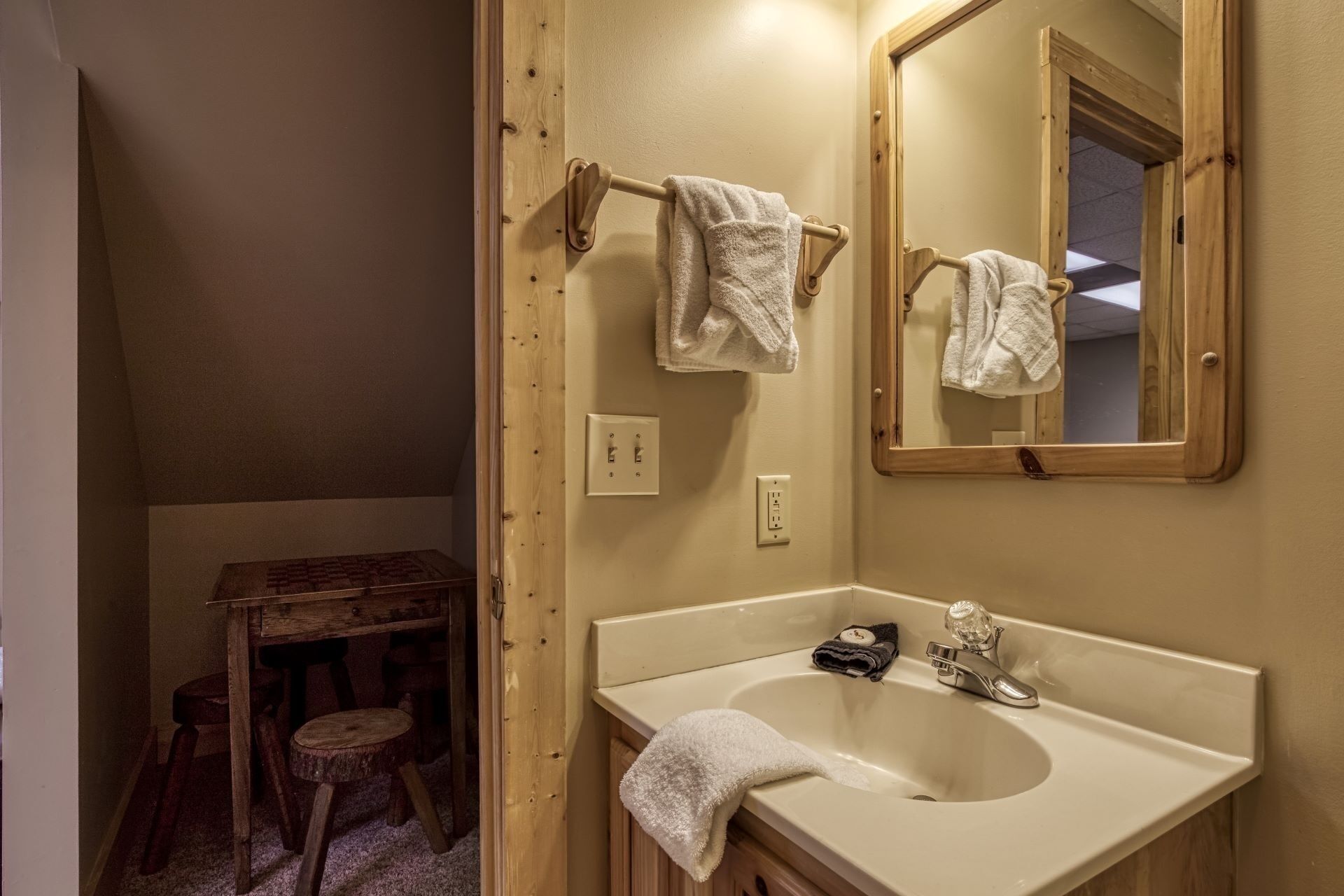 A bathroom with a wooden vanity, a sink, and a mirror. There are towels hanging on a towel rack and a stool near the vanity.