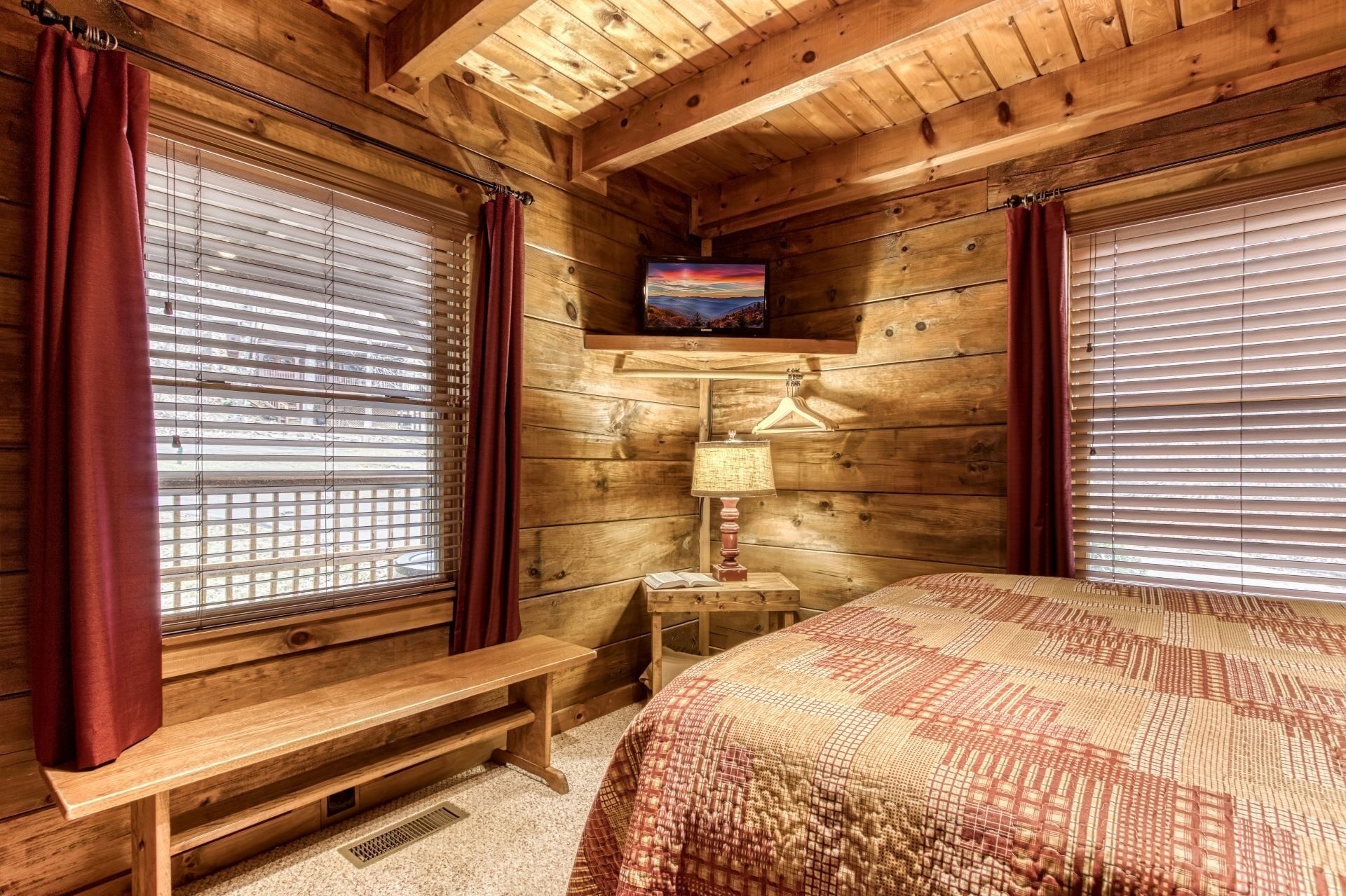 A cozy cabin room with wooden walls, a bed with a patterned blanket, a wooden bench, a table with a lamp, and a TV on the wall.