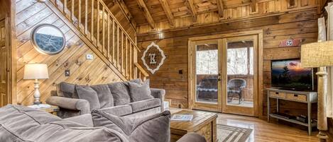 A cozy cabin interior with a wooden ceiling, a grey sofa, a television, and a staircase.