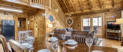 A wooden cabin interior with a dining table set for four, a sofa, a television, and a fireplace.