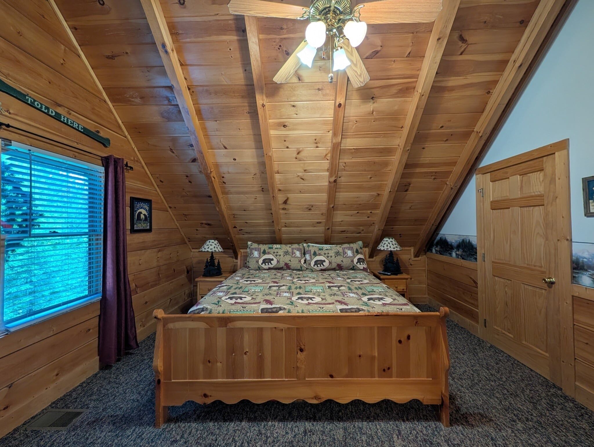 Un dormitorio de cabaña de madera con cama, dos mesitas de noche, ventilador de techo y ventana con persianas.
