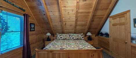 Un dormitorio de cabaña de madera con cama, dos mesitas de noche, ventilador de techo y ventana con persianas.
