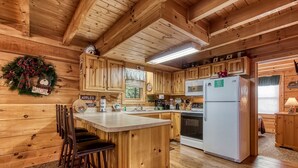 A wooden cabin kitchen with a central island, bar stools, wooden cabinets, a refrigerator, and a window with blinds. - ER245 - Almost Heaven Great Location - Close To Town! 3 Bedroom Cabin by RedAwning (Pigeon Forge)