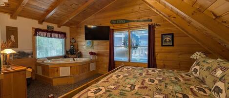 A cozy attic bedroom with a wooden ceiling, a large bed with a patterned quilt, a sitting area with a lamp, a bathtub, and a TV mounted on the wall.