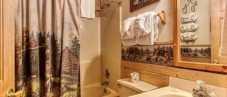 A bathroom with a shower curtain depicting a forest scene, a toilet, a bathtub, a sink, and a mirror with multiple towel hooks.