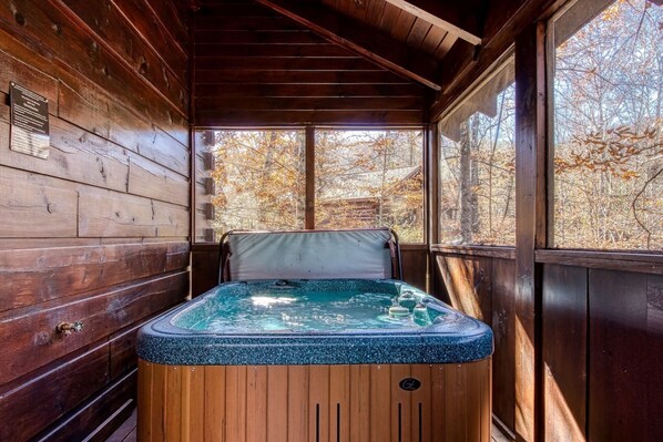 A wooden cabin with a hot tub in the center, surrounded by large windows offering a view of autumn trees.
