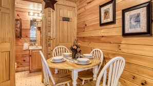 A wooden interior with a dining table set for six, surrounded by white chairs. A star decoration hangs from the ceiling, and framed artwork is displayed on the walls. - Er84 - Serenity Summit - Great Location. Close To All The Action! 1 Bedroom Cabin by RedAwning (Pigeon Forge)