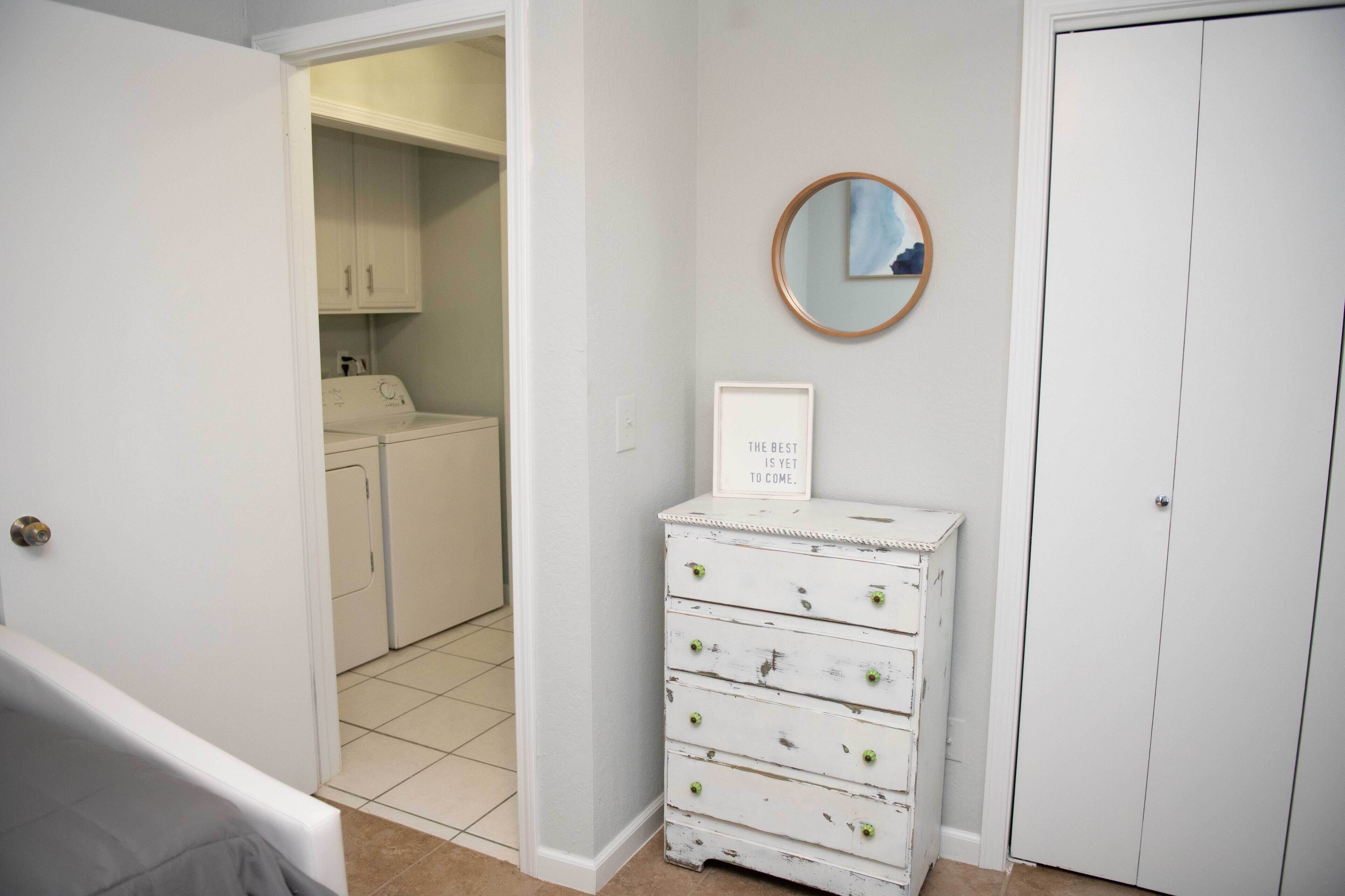Une buanderie équipée d’un lave-linge et d’un sèche-linge, une commode blanche avec des tiroirs, une citation encadrée accrochée au mur et une porte donnant sur une autre pièce.