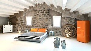 A bedroom with a stone wall, a bed, a dresser, a nightstand, a lamp, a vintage refrigerator, and a large wheel.