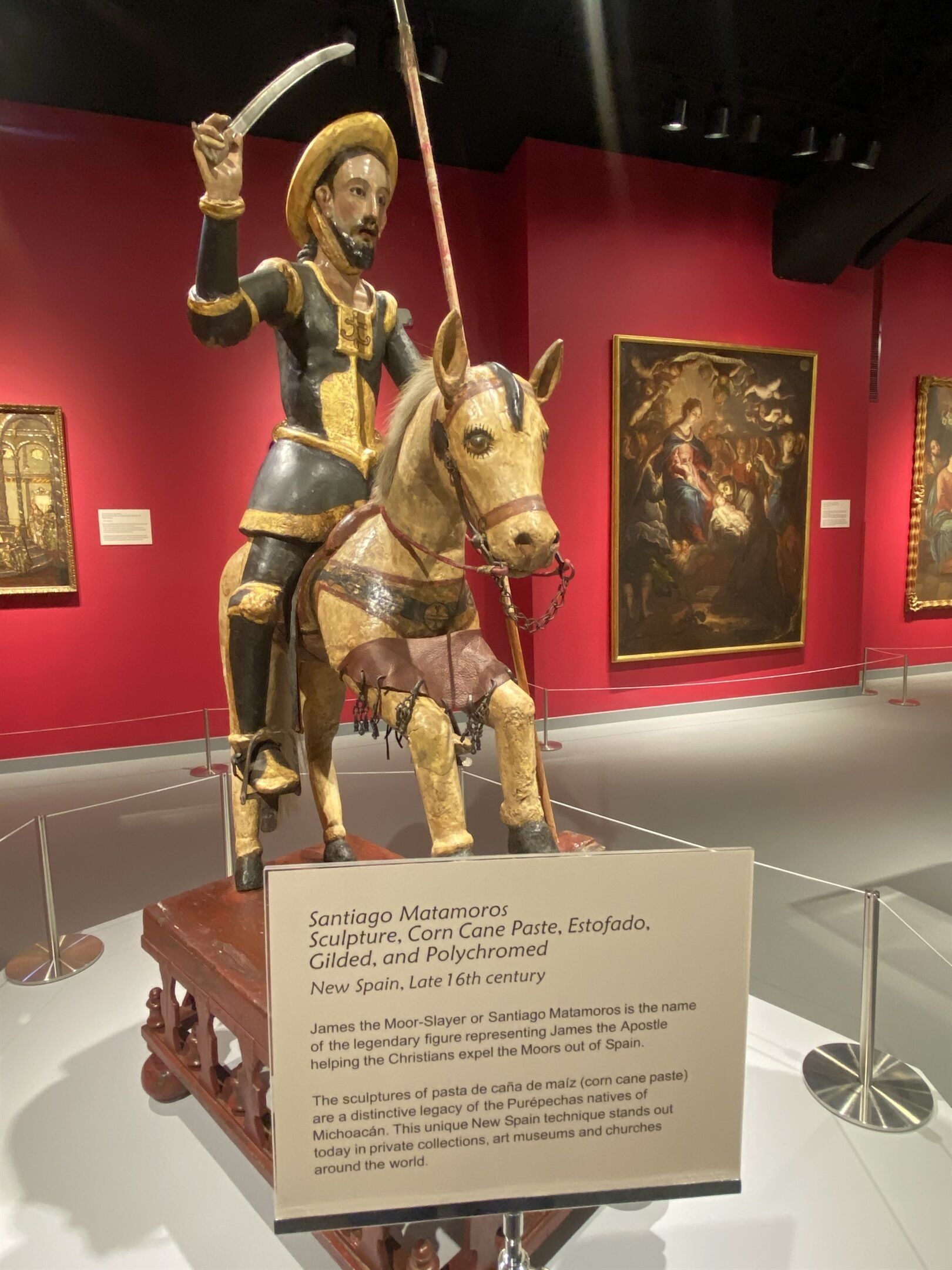 Une sculpture représentant Santiago Matamoros à cheval, exposée dans un musée aux murs rouges, avec des tableaux encadrés en arrière-plan.