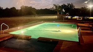 A swimming pool at dusk with surrounding deck chairs and a basketball hoop. - Happy Trails Cowboy Cabin HOT TUB POOL on 12 ac (Bandera)