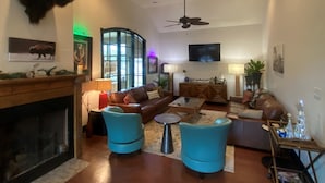 A spacious living room with a fireplace, a ceiling fan, and a large window. - Happy Trails Cowboy Cabin HOT TUB POOL on 12 ac (Bandera)