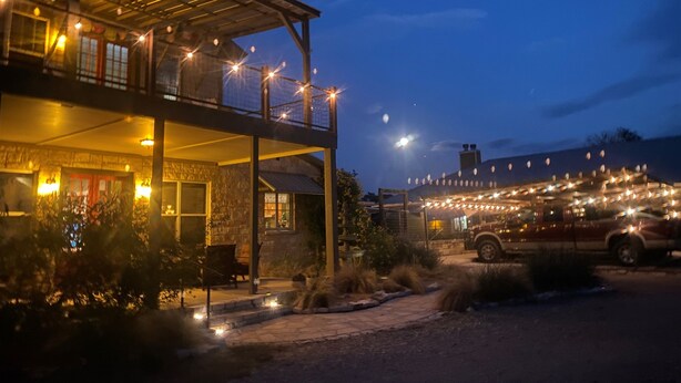 A two-story building with a covered outdoor seating area, a stone facade, and string lights. - Happy Trails Cowboy Cabin HOT TUB POOL on 12 ac (Bandera)